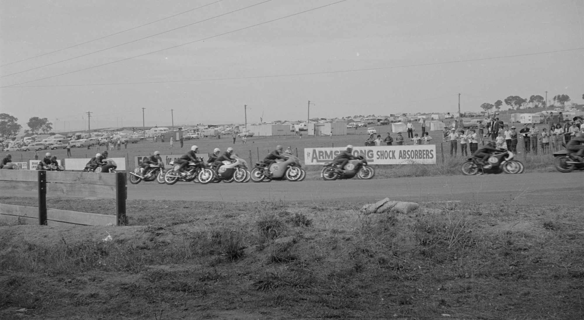 Black-and-white photograph of a tightly packed group of motorcycle racers leaning into a bend at Mount Panorama, Bathurst. Spectators watch from behind a wire fence and advertising hoardings, with rows of parked cars, caravans and tents covering the hillside beyond the circuit.